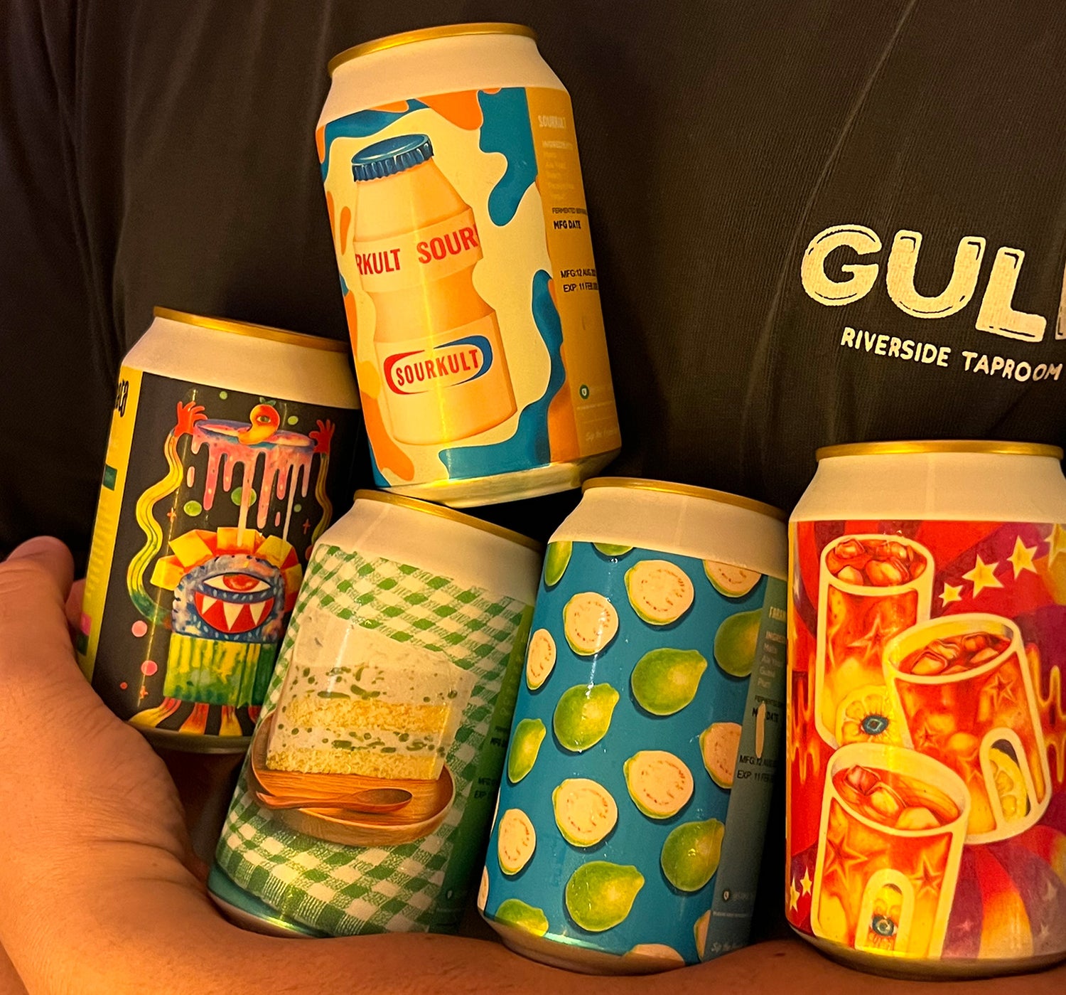 Five colorful beer cans held by a person wearing a black shirt with 'Gulp' branding.